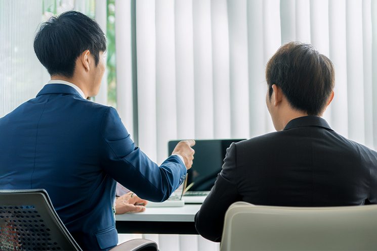 オフィスでPCを前に上司とやり取りするビジネスパーソン。口頭での瞬発的な会話には課題があっても、トラブル対応のメールのように状況・事実・原因・対策を論理的に構造化して伝える「非同期テキストコミュニケーション」において、エンジニアが独自の強みを発揮できる場面を象徴している。