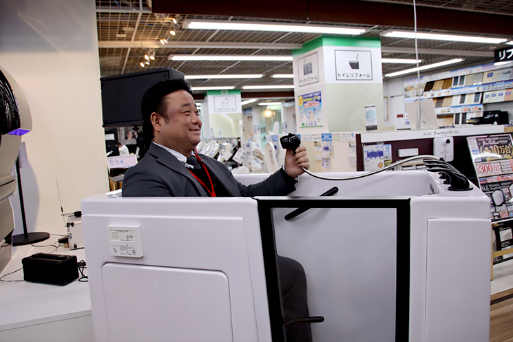 ヤマダデンキの社員が「ミライ人間洗濯機 社会実装版」の中に座り、実際の入浴姿勢を実演している様子。足の不自由な方でも車椅子から最小限の動作で移乗できるよう設計された筐体のサイズ感と、145万円（税別）という価格で介護現場や一般家庭への普及を目指す製品の実用性が示されている