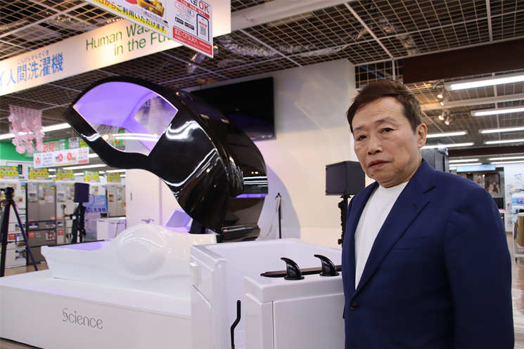 ヤマダホールディングス顧問の笹澤淳氏が、共同開発したサイエンス社の「ミライ人間洗濯機 社会実装版（145万円）」の横に立ち、普及への想いを語る様子。背景には大阪・関西万博で話題となった6,000万円のハイエンドモデルが展示されており、介護現場や一般家庭への導入を見据えた戦略的な製品ラインナップの対比が示されている