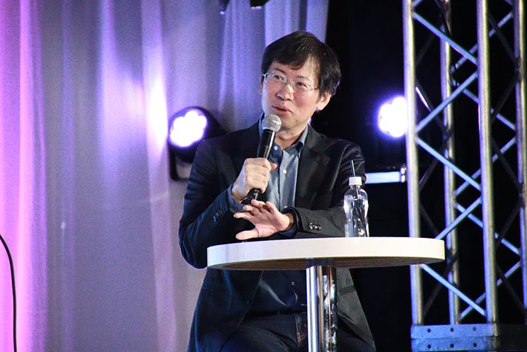 『Eight EXPO 2026』の特別講演に登壇する、日立製作所 CAXO（Chief AI Transformation Officer）の吉田順氏。 マイクを握り、会場に向けて語りかけている。電力や鉄道などのインフラ現場における「熟練者の暗黙知」をいかにAIに継承するかという課題について説いている。