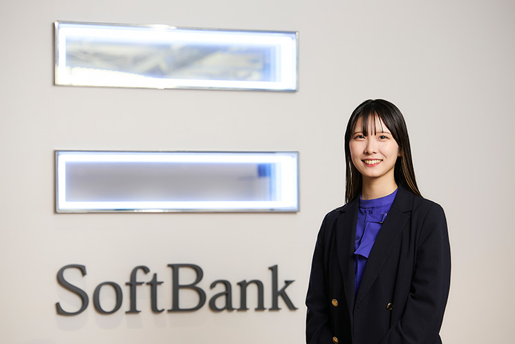 ソフトバンクの木口佳南さん。ネイビーのジャケットとブルーのブラウスを着用し、カメラに向かって微笑んでいる。背景の白い壁には、ソフトバンクのロゴがはっきりと表示されている