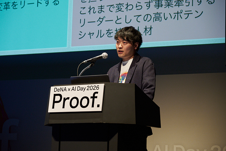 「DeNA × AI Day 2026」の壇上でプレゼンテーションを行う採用戦略部部長の石井敬時氏。「Proof.」と記された演台に立ち、背後のスクリーンには採用基準の変革に関する資料が映し出されている。数万件の採用データを学習したAIの判断精度がベテラン選考官と同等であることを実証し、選考プロセスにおけるAI活用を「当たり前の前提」とする同社の先進的な姿勢を象徴する登壇シーン。