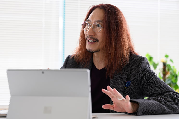 デスクでノートPC（Surface）を前にし、真剣な眼差しで「観察」の重要性を説く澤円氏。前職の経験に固執する「出羽守」になることを戒め、まずは新しい職場の「暗黙の了解（コンテキスト）」を読み解くために徹底的に周囲を観察するというマインドセットへの転換を促す文脈で使用されています。