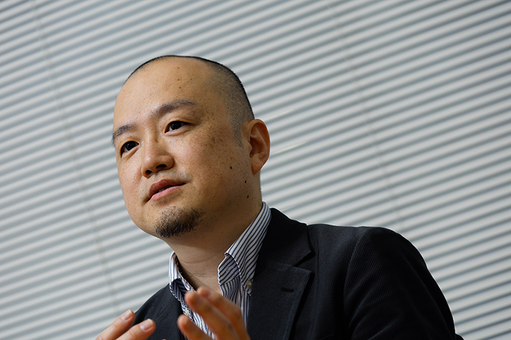 真剣な表情で語る間野健介氏。俳優時代の「役作り」において、戦時中の人々を理解するために自ら都内の防空壕へ足を運び、暗闇や湿り気を肌で感じた経験を回想している。知識としての理解を超えた、身体感覚を伴う「想像力」こそが、エンジニアに求められるユーザー理解の原点であると説く一場面。