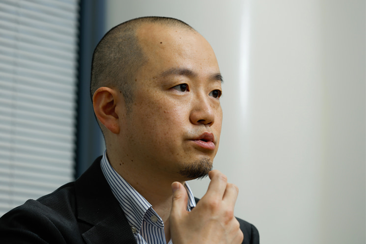 考え込む表情の間野健介氏。経験を積むほど強くなる「思い込み（バイアス）」を排除するため、現場での「キャリブレーション（補正）」の重要性を語る場面。直射日光下で見えなくなるタッチパネルの例を挙げ、画面越しでは得られない「情報の解像度」を物理的な検証によって高めることが、設計バグを防ぐ鍵であると提示している。