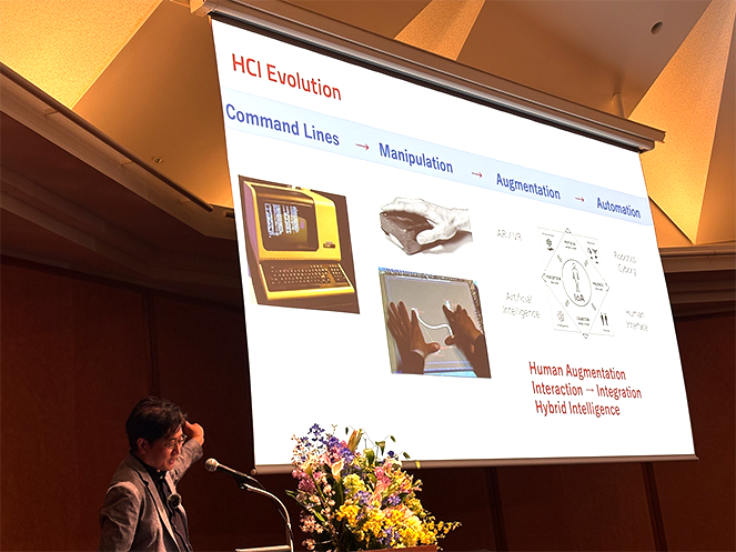 東京大学の最終講義にて、暦本純一教授がHCI（人間・コンピュータ・相互作用）の進化を解説する様子。1970年代のビデオ表示端末VT100から、マウスによる直接操作、AR/VRによる人間拡張、そしてAIとの統合（Integration）へと至る歴史が投影されており、UIが再びテキスト入力へと「先祖返り」している現状の違和感を提示する文脈で使用されている。