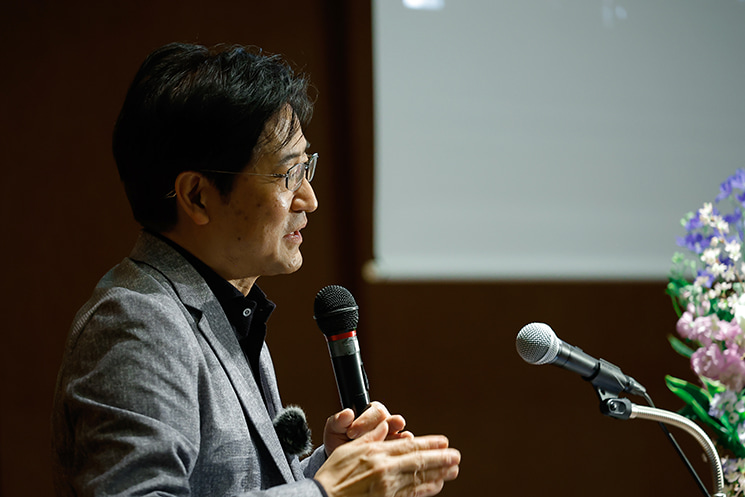 2026年2月の最終講義でマイクを手に語る暦本純一教授。ChatGPTやClaudeの普及により、UIが再びテキスト入力（チャット欄）へと「先祖返り」している現状に対し、それが真の進化なのか、あるいは次のサイクルへの予兆なのかを聴衆に問いかけている。エンジニアが直面する、AI時代のインターフェースにおける不自然さと課題意識を象徴する一場面。