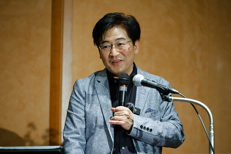 東京大学最終講義の壇上で、UIの歴史的変遷と「現時点での結論」を語る暦本純一教授。1970年代のビデオ表示端末「VT100」から、マウスによる「直接操作（ダイレクト・マニピュレーション）」、そしてテクノロジーを身体化する「人間拡張」へと至る進化のプロセスを解説し、現代のチャットUIへの回帰に対する課題意識を提示している場面。