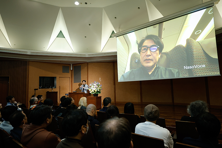 暦本純一教授の最終講義にて、自身の研究プロジェクト「SottoVoce / NasoVoce」を紹介する様子。スクリーンには新幹線内の騒音環境下でスマートグラスを着用し、口パク（サイレントスピーチ）によってAIと意思疎通を図る実証実験の様子が映し出されている。周囲に迷惑をかけず、思考をテレパシーのようにAIへ伝達する未来のインターフェースを象徴する資料。