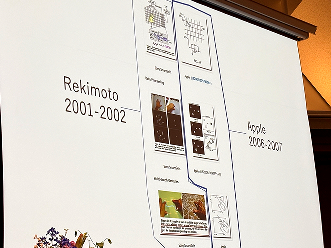 iPhoneのマルチタッチ特許を巡る裁判で比較された資料のスライド。左側には2001年に暦本純一教授が発表した「SmartSkin」の論文図解、右側には2006年にApple社が主張した図面が並ぶ。判決によりAppleの主張が退けられ、暦本氏の研究が現代のスマートフォン操作の基礎となった歴史的事実を示す、技術史において極めて重要な証拠写真。