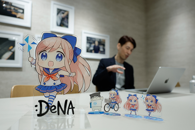 手前にDeNAの「Mobage」公式キャラクターなどのアクリルスタンドを配し、奥にfreee株式会社CISOの茂岩祐樹氏が佇むインタビューの結びカット。技術がどれほど進化しても変わらない「動くものを作るのは容易だが、動き続けさせることは困難で価値がある」というエンジニアリングの原理原則と、自分の技術で誰かの日常を守り続ける執念の先にある醍醐味を語るシーン。