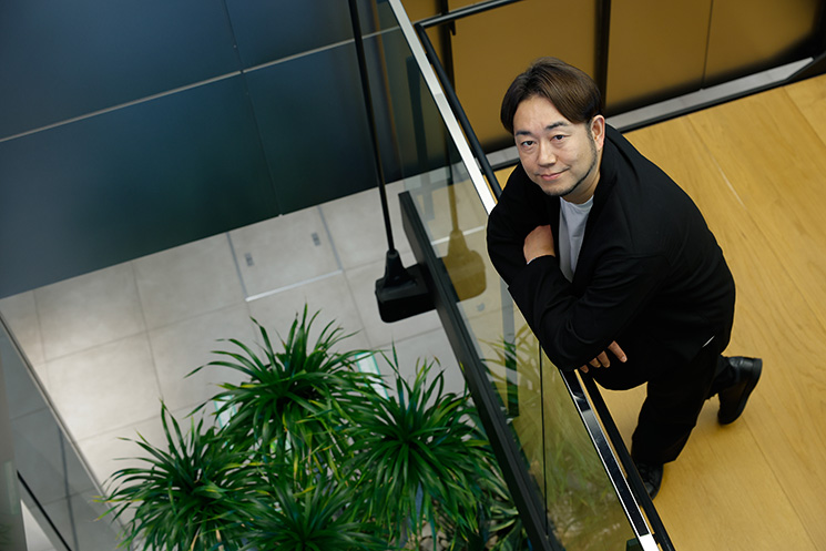 MIXIのオフィスにて、手すりに寄りかかり俯瞰の構図でカメラを見つめる竹野太一さん