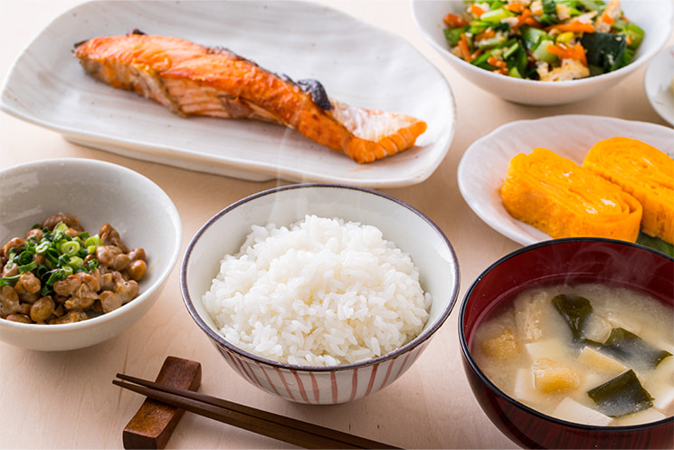 白米、味噌汁、焼き鮭、卵焼き、納豆、和え物がきれいに盛り付けられた一汁三菜の日本の伝統的な朝食。この記事において、単に栄養（正論）を摂取できれば形は何でもいいという「ミキサー飯理論」の対比として提示されており、ビジネスの指導場面において「相手が受け取りやすい言葉の盛り付けや配慮」がいかに重要であるかを視覚的に伝える役割を果たしている