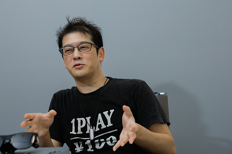 「1PLAY ￥100」と書かれたTシャツを着用し、インタビューで身振り手振りを交えながら熱弁するナル先生（GOROman）のポートレート写真。AIが自律的に動きコードを生成する時代において、構文やAPIの知識は化石となり腐るため、エンジニアに必要なのはシステムを自ら作る「野生の衝動」であるという主張について書かれている。