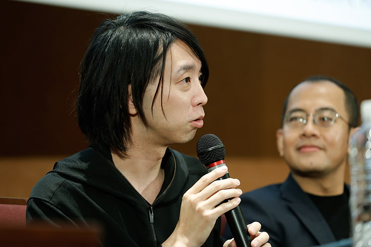 暦本純一教授の最終講義にて、学生時代に教授がコーヒーを淹れに来るわずか45秒の間に「これこうしたら面白いんじゃない？」と一言残して去っていった日常を振り返り、新しい研究をひらめくためにはそれほど「心が軽い人」でいることが重要であると、後ろで見守るAdiyan Mujibiya氏と共にマイクを手に語る落合陽一氏