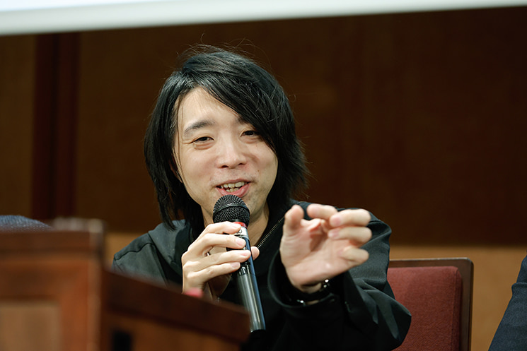 暦本純一教授の最終講義にて、東京大学のプレハブ校舎「暫定アネックス」に研究室があった初期時代を振り返り、物理的に建物が揺れて顕微鏡のピントが合わなかったり台風で潰れそうになったりしたカオスな開発環境と、その不確実性（揺らぎ）がタフな研究力を育てたエピソードを、手振りを交えて笑顔で語る落合陽一氏