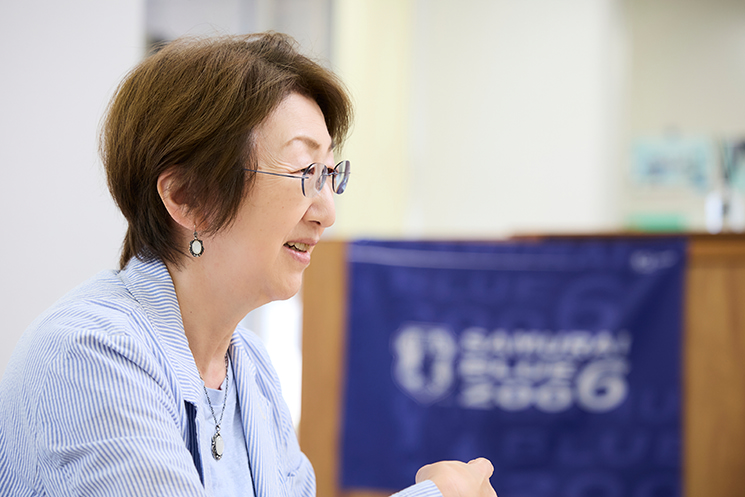「SAMURAI BLUE」のフラッグを背景にインタビューに答えるつくば言語技術教育研究所所長・三森ゆりか（さんもり・ゆりか）氏。元日本サッカー協会会長の田嶋幸三氏がドイツ・ケルン体育大学留学中に現地の子どもたちが論理的に説明し合う姿に衝撃を受け、日本のスポーツ界やビジネス界における「理屈で考え、組織で言語化する力」の必要性にいち早く気づいたエピソードを語る文脈を示している