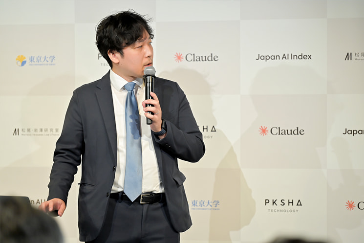 2026年6月の「Japan AI Index」発表会にてマイクを持ちスピーチする東京大学の岩澤有祐准教授。第1回レポートおよびダッシュボードの同時ローンチを2026年秋（10〜11月頃）に予定しているスケジュールを明かし、その後は四半期に1回程度のペースで継続的なデータアップデートを行い、指標の鮮度を維持していく中長期ロードマップの方針を語っている