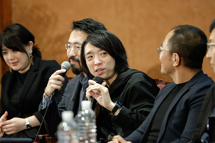 暦本純一教授の最終講義にて、杜氏がお酒（酵母）を無理に作らせるのではなく発酵しやすい環境を整えるように、AIが自律的に動く環境をエンジニアが設計する「発酵」のような開発アプローチの重要性について、隣り合う玉城絵美氏、石黒祥生氏、Adiyan Mujibiya氏らに囲まれながらマイクを手に語る落合陽一氏