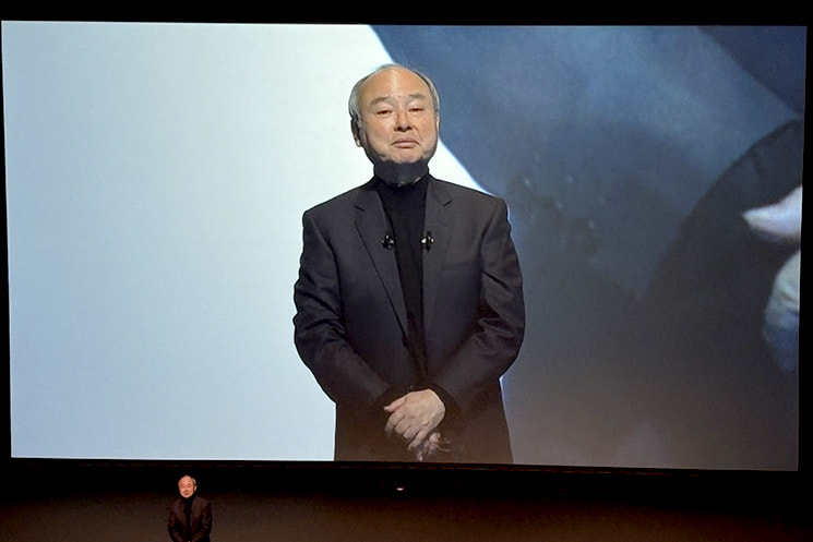 第46回ソフトバンクグループ定時株主総会（2026年）にて、大画面スクリーンに映し出された自身の姿を背に、ユーモアを交えて語る孫正義氏。株主価値1,000兆円の宣言が決して冗談や思いつきではなく、外れれば非難を浴びるリスクを背負いながらも、言ったことは必ずやり遂げるという経営者としての覚悟を示す場面