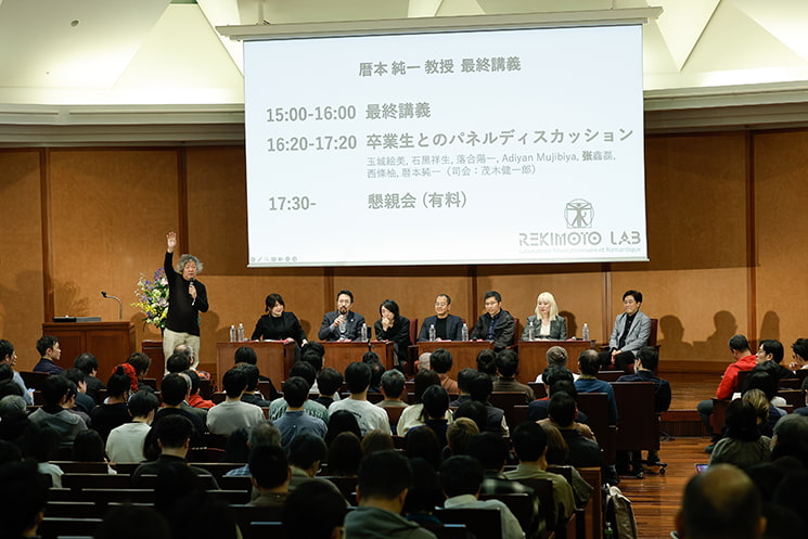 東京大学の伊藤謝恩ホールにて、満員のオーディエンス席からステージを一望した風景。巨大スクリーンに「暦本純一教授 最終講義」「卒業生とのパネルディスカッション」のプログラムが投影される中、司会の茂木健一郎氏が手を挙げながら会場からの「研究や開発の原点」に関する質問をファシリテーションし、暦本教授や卒業生パネリストたちが耳を傾けている質疑応答の様子