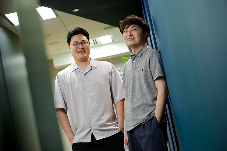 アクストリア株式会社のエンジニア、奥藤陸矢さん（左）と小林勇輝さん（右）のツーショット写真。オフィスの廊下で笑顔で立っている様子