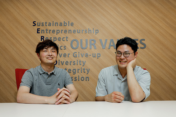 アクストリア株式会社の「OUR VALUES」が掲げられたオフィスで笑顔を見せる小林勇輝さん（左）と奥藤陸矢さん（右）。受託開発・SES・自社プロダクト開発におけるエンジニア採用や今後の展望をアピールする締めくくりのツーショット写真