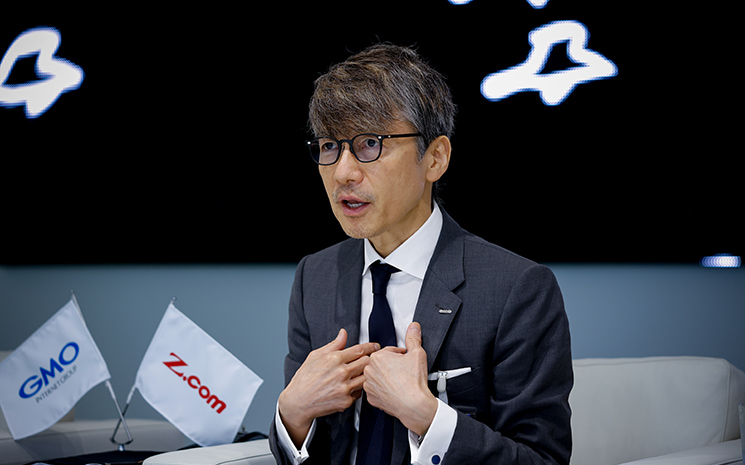 20代前半の苦悶した時期を振り返り、環境や他人のせいにすることをやめて「すべての責任は自分にある」という自責思考へ切り替えた覚悟の瞬間を、自身の胸に手を当てながら真剣な表情で語るGMOの熊谷正寿氏。このマインド転換こそが、後のGMOインターネットグループ創業の原点になったと説明するシーン