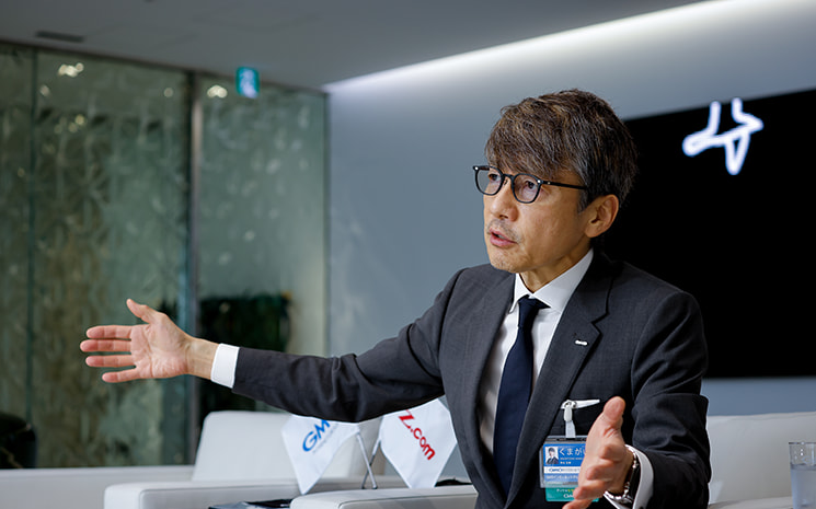 GMOの熊谷正寿氏が、生成AI（Claude）を用いた開発において、キーボードの代わりに音声入力を活用する生産性の高さについて身振り手振りを交えて熱弁する様子。自身の体験に基づき、グループ全社8000人にグースネックマイクを導入し、音声入力を徹底させるプロジェクトを即座にスタートさせた背景を語るシーン