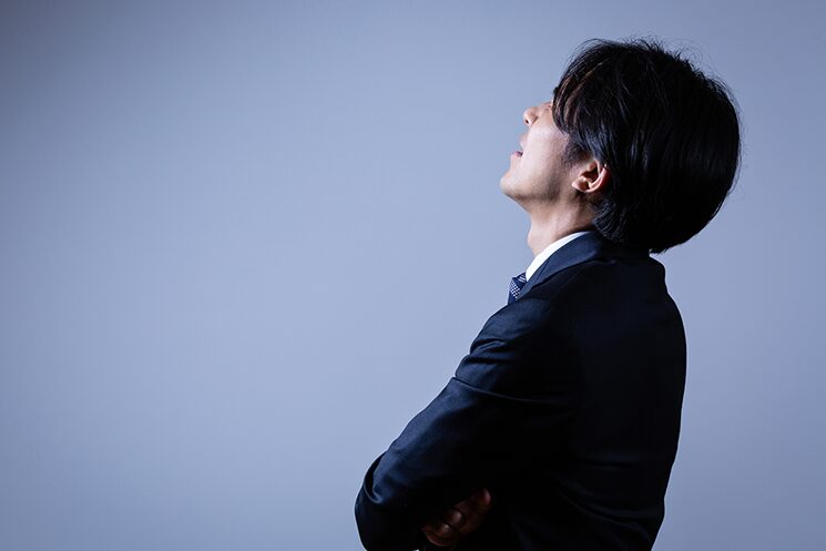暗い背景の中、腕を組みながら真剣かつ戸惑った表情で、見えない何かを見上げている40代エンジニア風の男性。これは、好条件のスカウトを前に「新天地で通用するか」と足がすくむ相談者の葛藤を視覚化したもの。澤円氏が勧める「情報のデバッグ」の必要性を象徴するイメージ画像