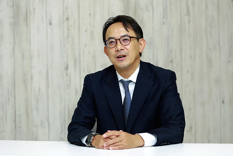 眼鏡をかけ、紺色のスーツに身を包んだ前LINEヤフー代表取締役会長の川邊健太郎氏が、真剣な表情で前方を見据えるポートレート。記事本文では、AIがどれだけ進化しても究極の「指示待ち」であり、人間の能動的な指示がなければビジネスは始まらないと主張するセクションに配置されている
