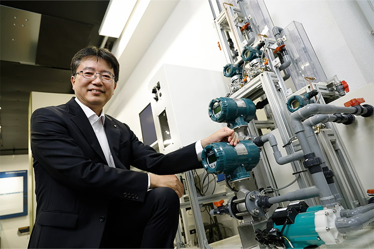 製造設備の前に立つ横河デジタル代表取締役社長・鹿子木宏明氏。化学プラントの制御にAIを適用してきた経験を踏まえ、AI時代の技術者には技術を極めるだけでなく、「ビジネスになる技術」を見極める力が求められると語る
