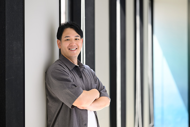 アワーズシップ株式会社の代表取締役・塩田悠麻さん。窓際に立ち、腕を組んで笑顔の一枚