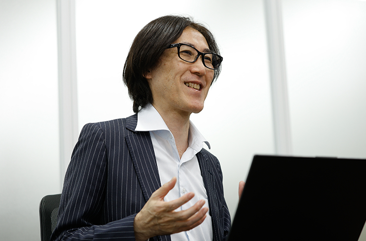 秋葉原で購入した実機機器などを自宅に置き常に学び直す姿勢を持ち続ける小原氏。新しい分野では誰もが初心者であるとし、自ら仮説を立てて考え続け試行錯誤する探究心がエンジニアの成長に不可欠であることを笑顔で語る様子